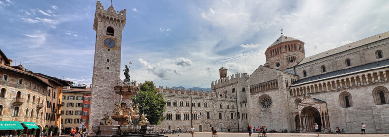 Piazza Duomo in Trento. In the background, the Civic Tower, Palazzo Pretorio and San Vigilio Cathedral, the city's cathedral. In the centre, the Neptune fountain. Behind the fountain stands the only tree in the square: a lime tree. The square, in the image, is lived in. There are some people sitting on the steps of the fountain, someone is taking photos and a small group is posing in front of the cathedral to have their picture taken. Everyone is wearing summer clothes. The blue sky is streaked with the white of light clouds.